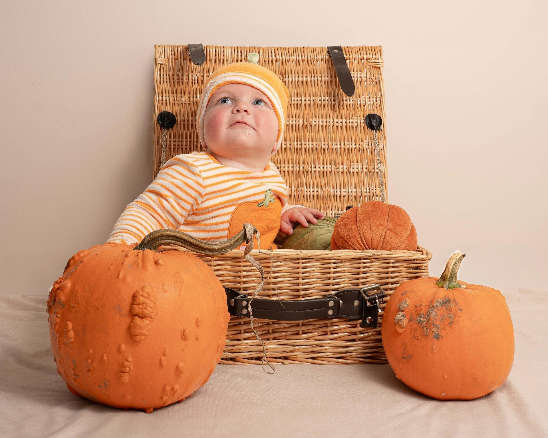 Baby sits in a wicker hamper between pumpkins