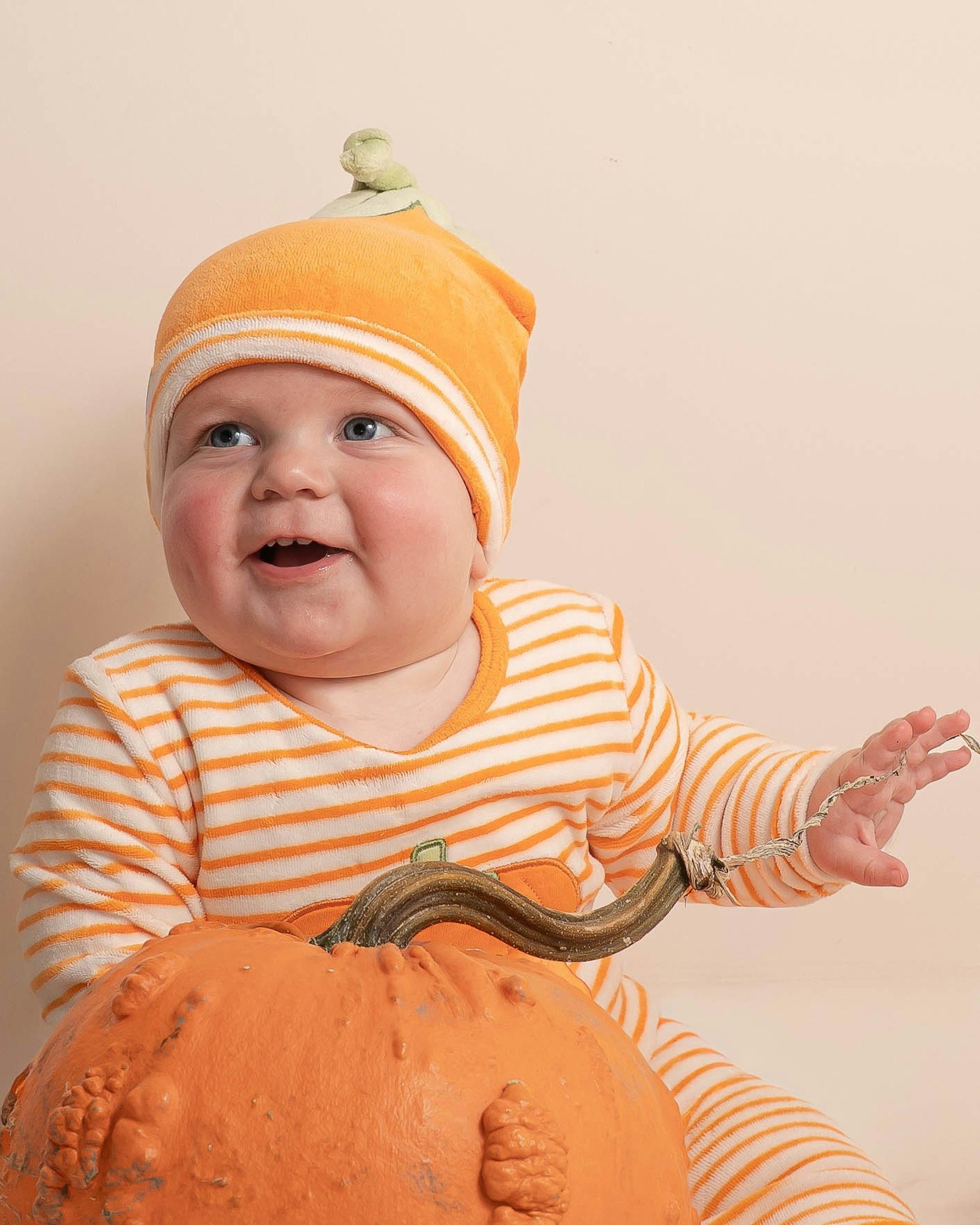 Baby in an orange pumpkin hat laughing beside a pumpkin