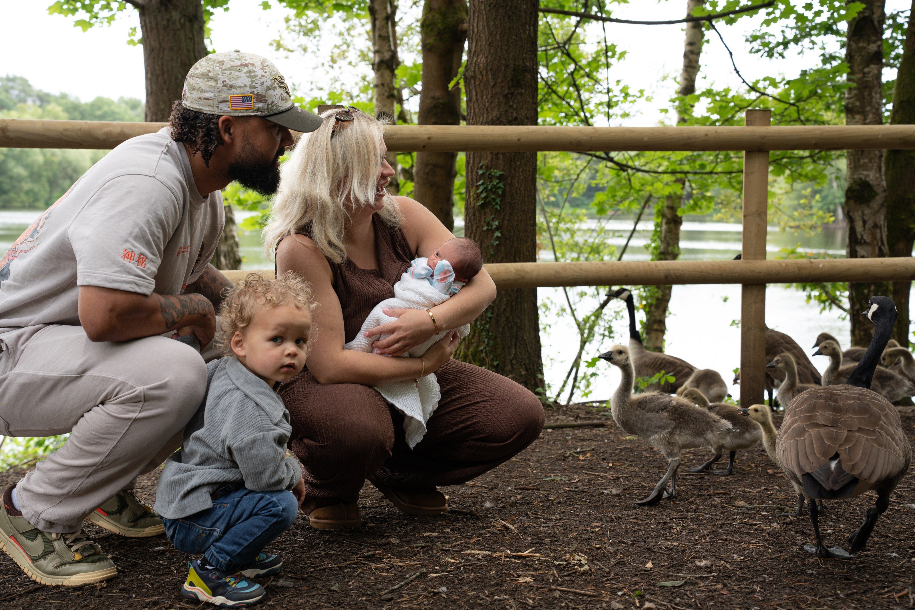 Mum cradles the baby while dad and toddler feed the geese by the lake