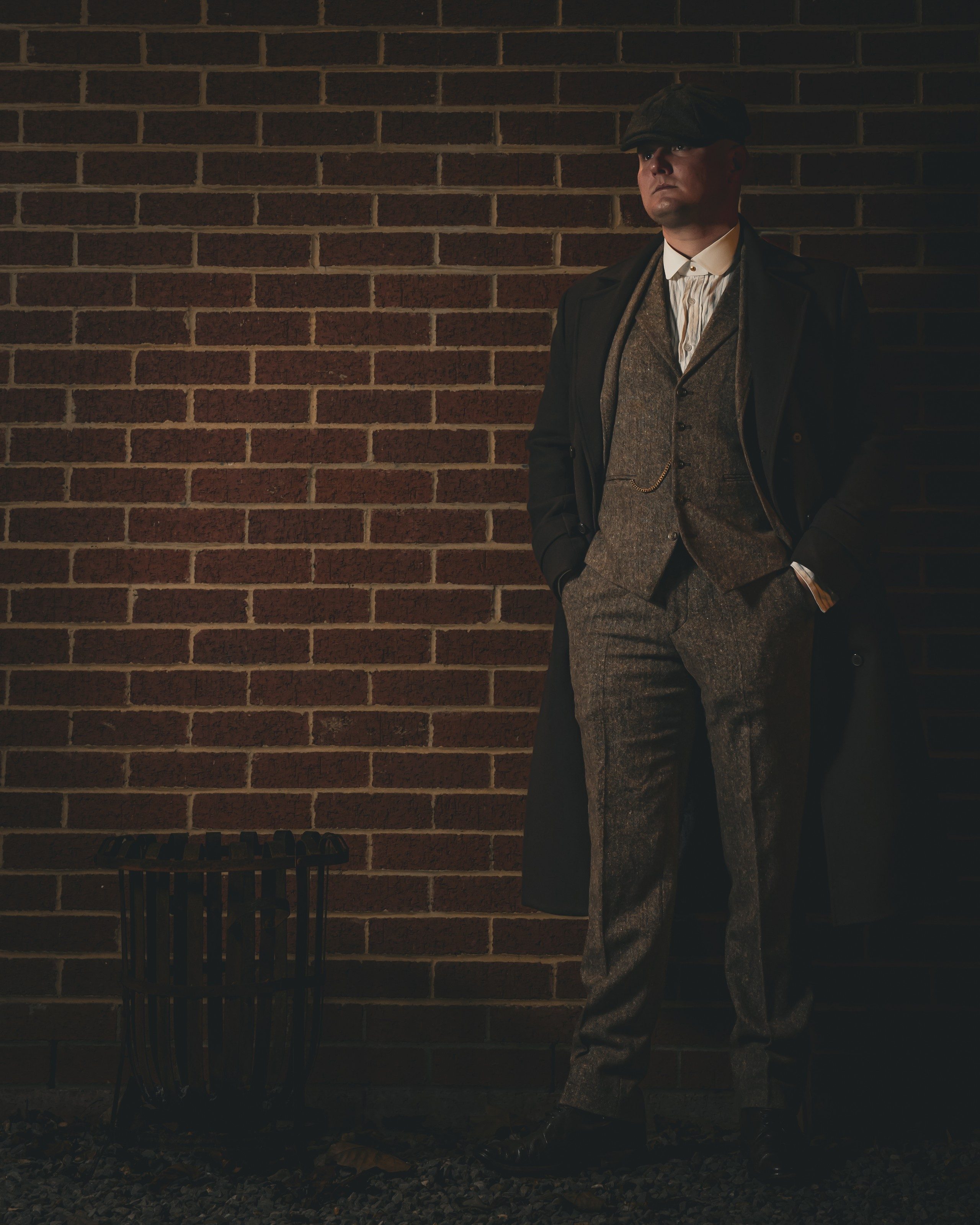 Man in a flat cap and tweed suit beside a brick wall at night
