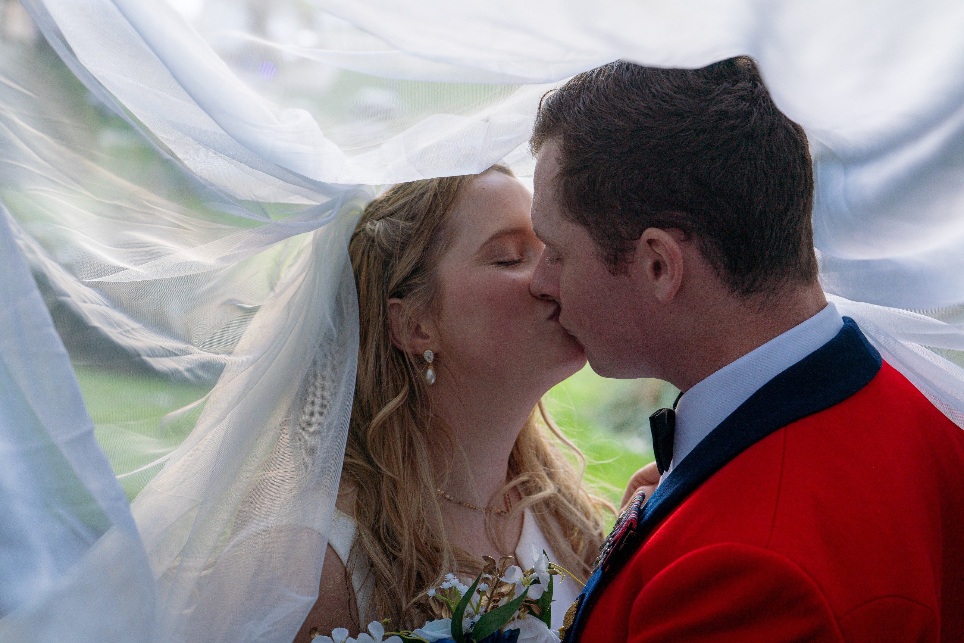 Groom in red military mess dress kisses his bride beneath her veil