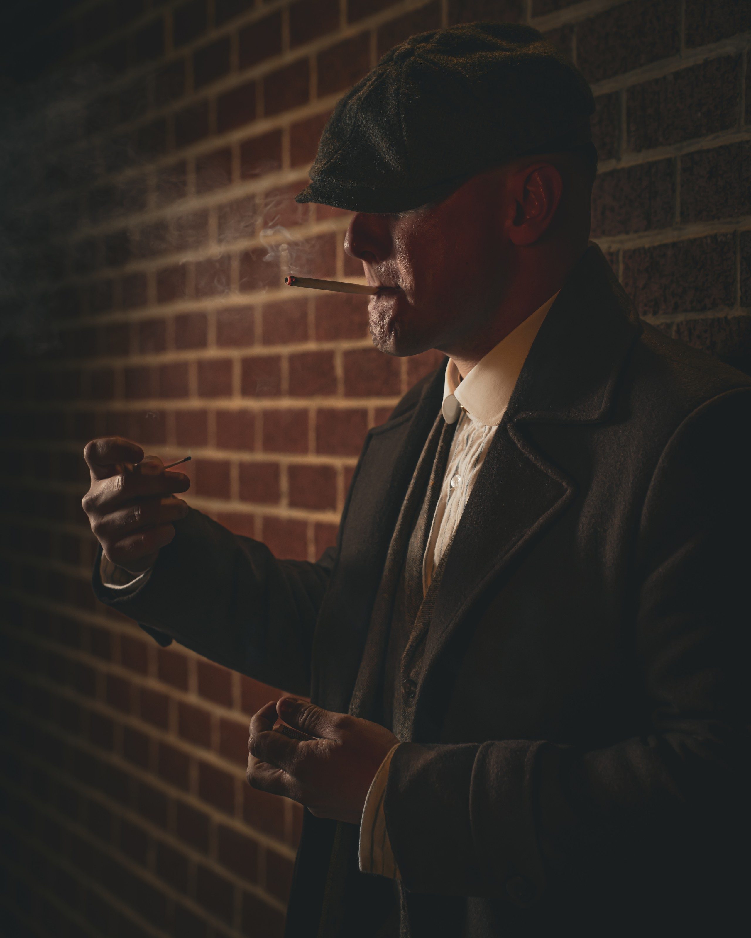 Man in a flat cap lights a cigar against a brick wall