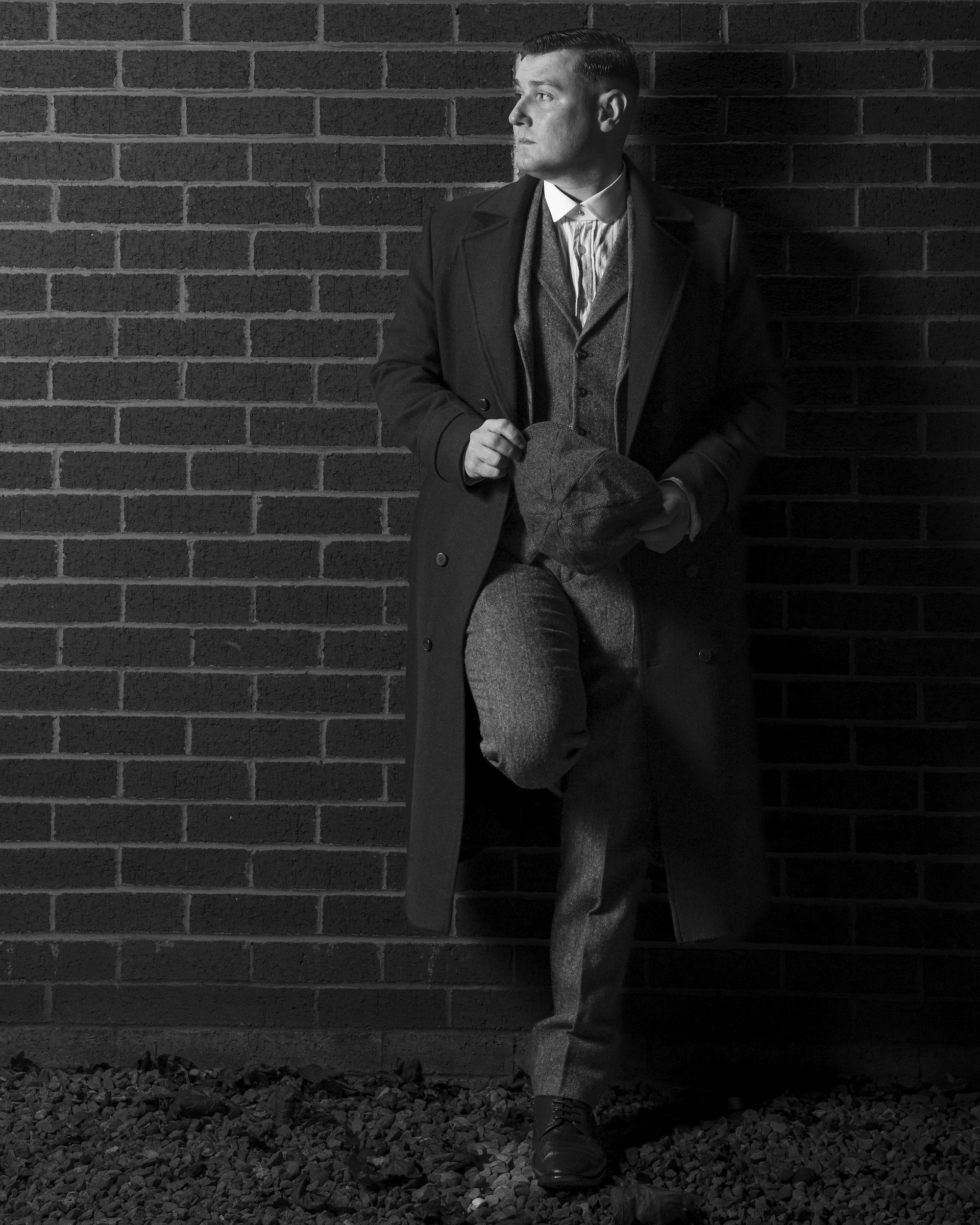 Man in an overcoat leans against a brick wall, in black and white