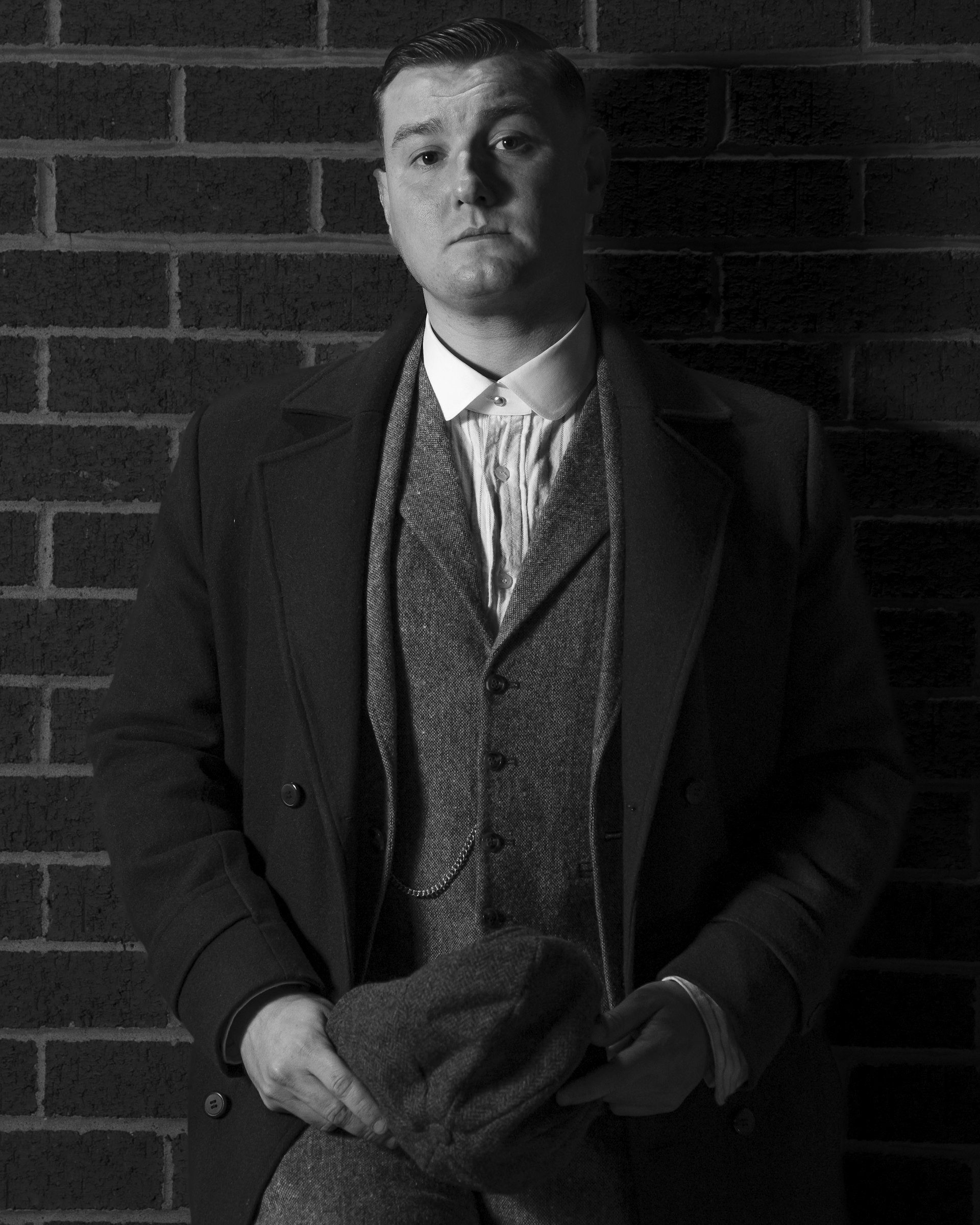 Man in a waistcoat and overcoat sits against a brick wall, in black and white