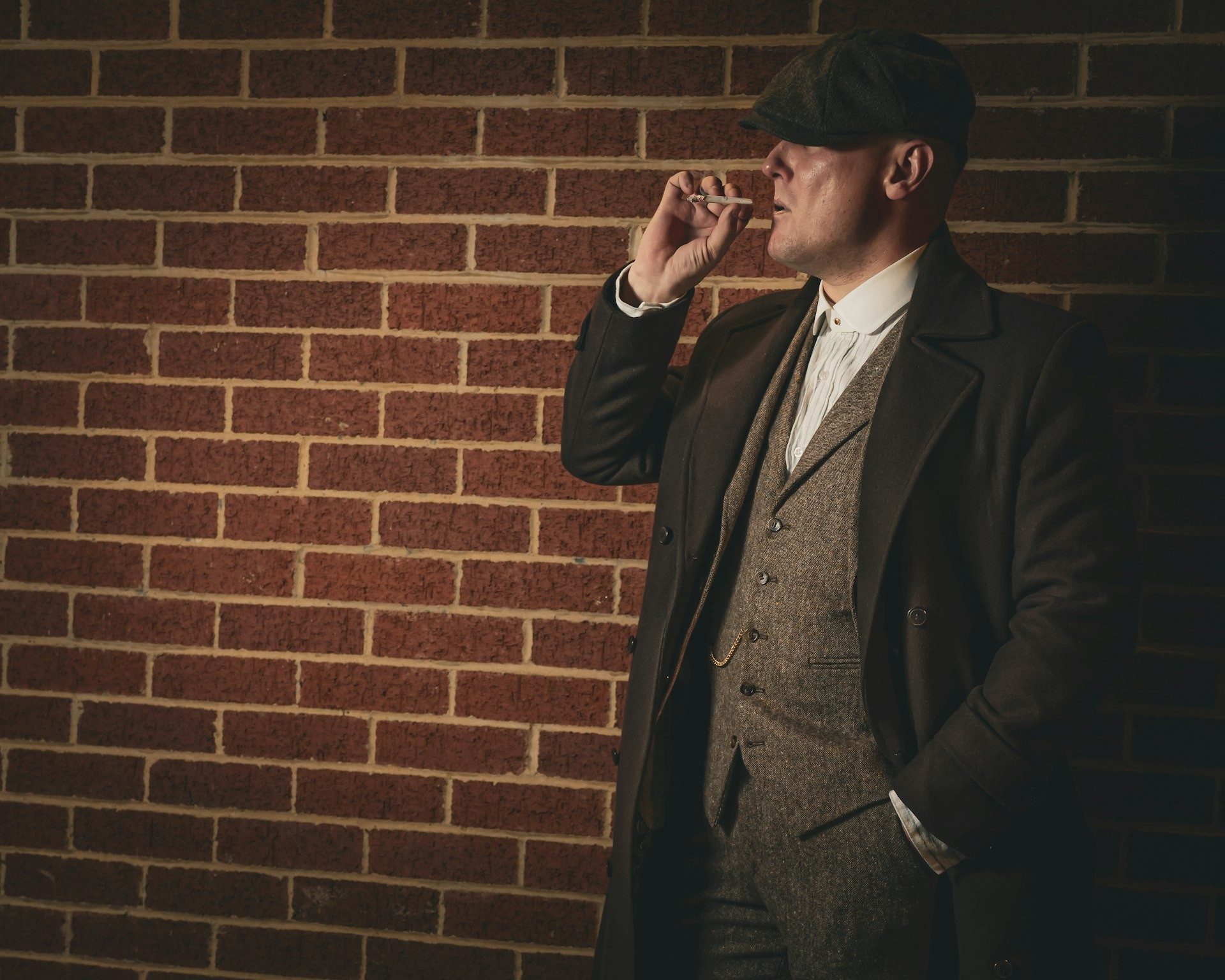 Man in a flat cap smokes against a brick wall