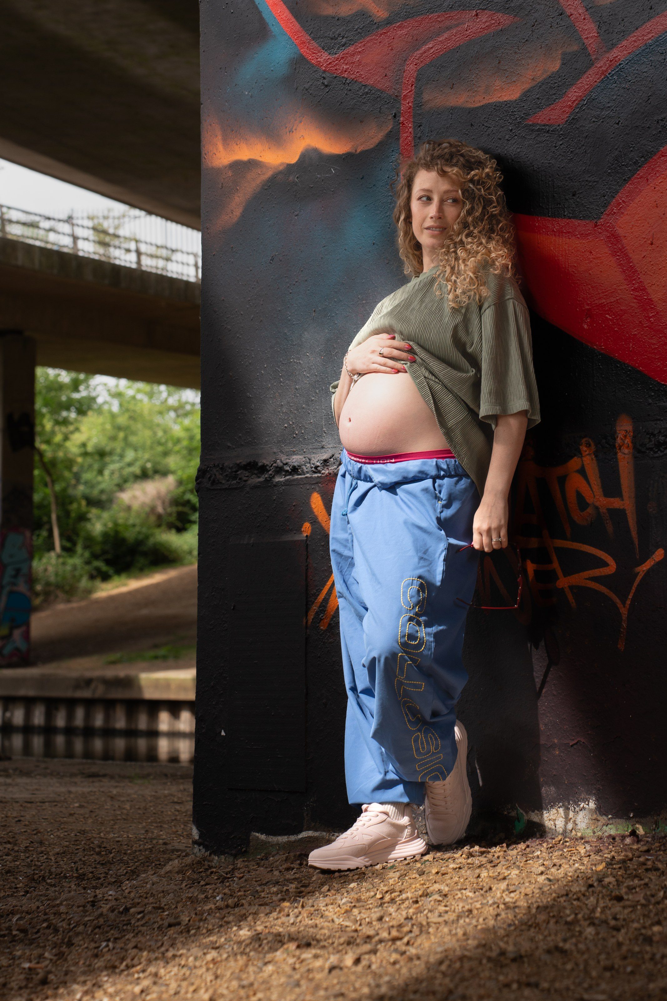 Model in blue trousers leans against a graffiti pillar under the bridge