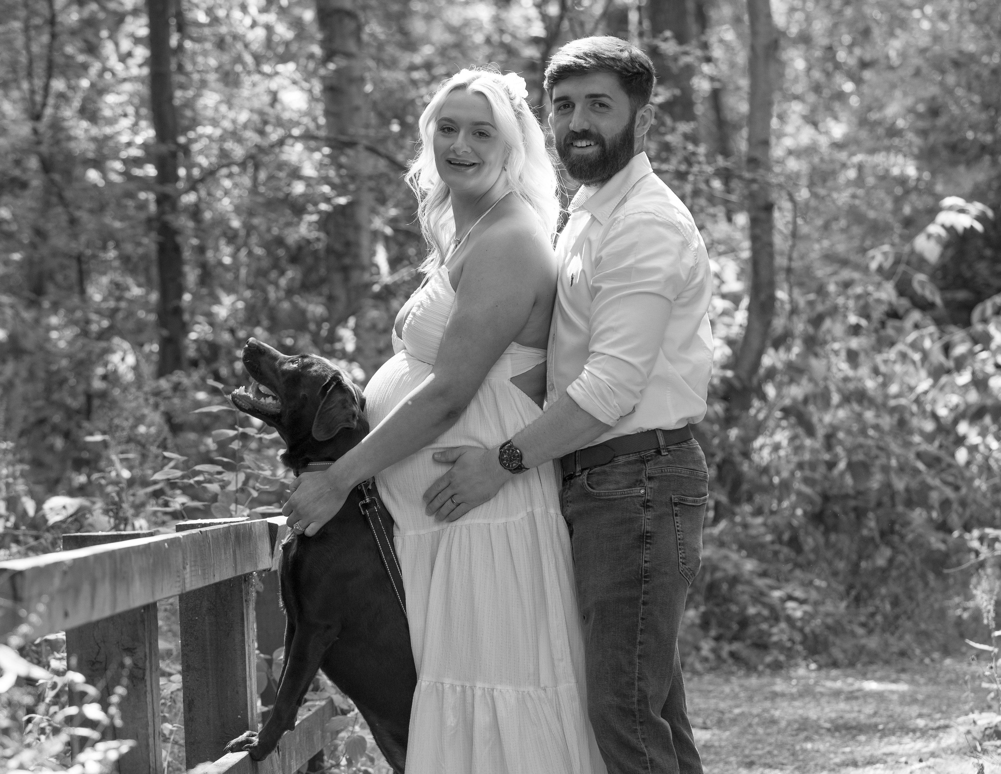 Expectant couple and their dog on a woodland path, in black and white